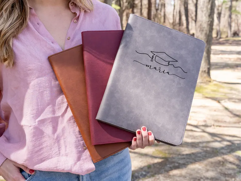 A woman holding three engraved leather portfolios in tan, burgundy, and gray with a graduation cap design, showcasing practical and stylish graduation gifts she can use in her career. Etsy