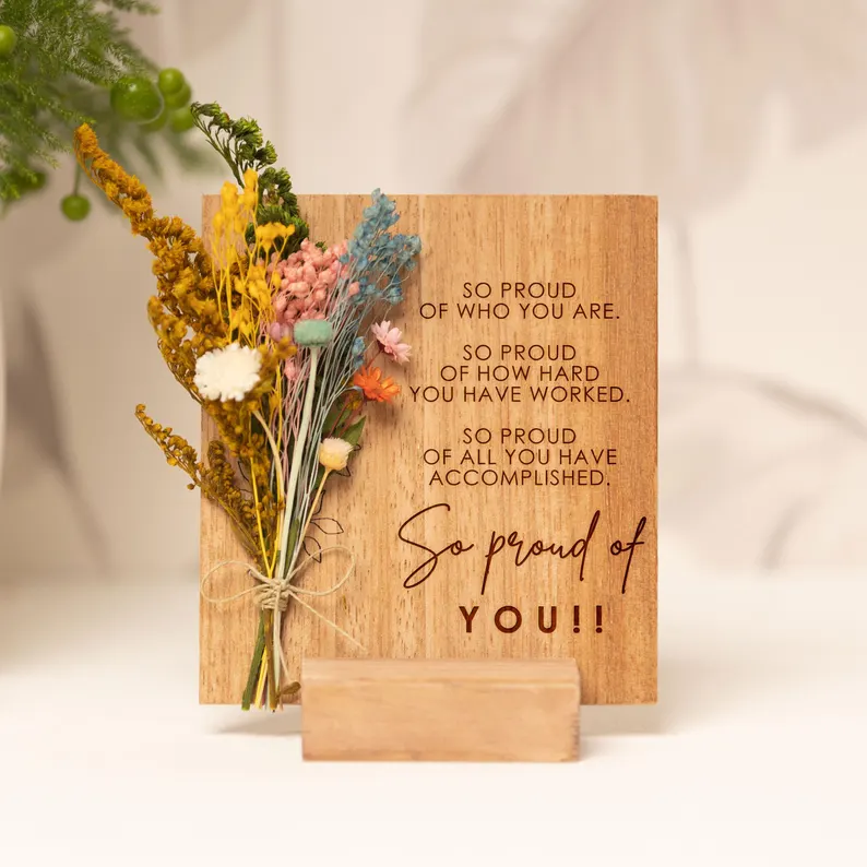 Wooden sign engraved with "So Proud of You" paired with a colorful dried flower bouquet on a stand, among the most heartfelt graduation gifts. Etsy