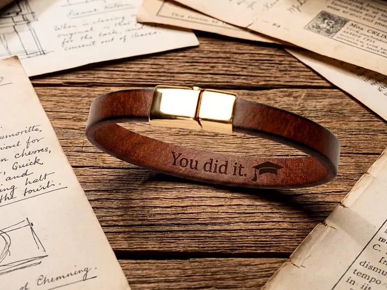 Brown leather bracelet engraved with "You did it" and a graduation cap icon, displayed on vintage papers, among the most meaningful graduation gifts. Etsy