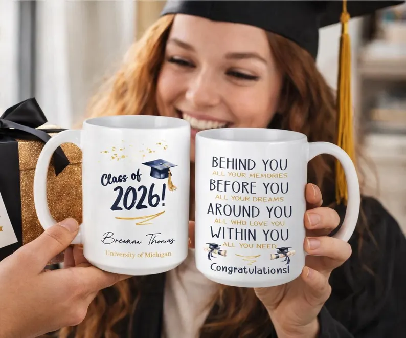 Smiling female graduate in cap holding two white personalized mugs printed with "Class of 2026" and an inspirational quote, among the most thoughtful graduation gifts. Etsy