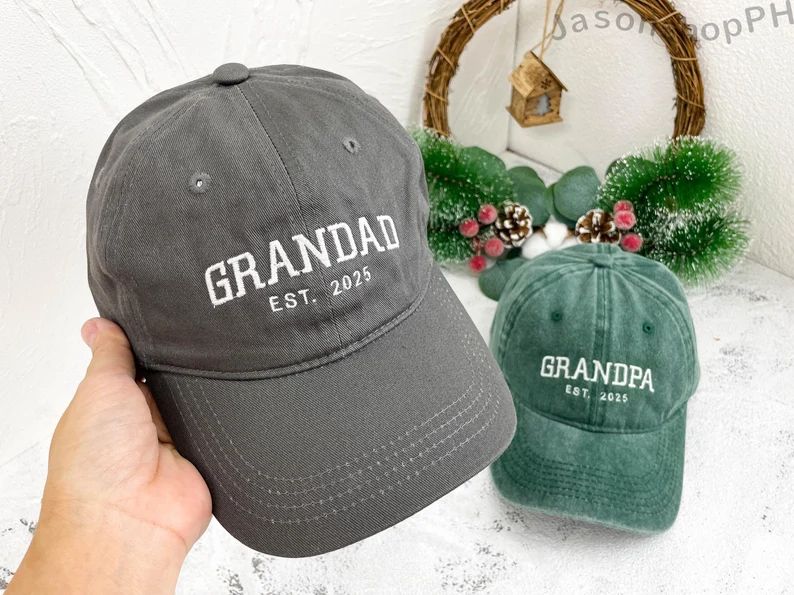 A hand holding a charcoal embroidered "Grandad Est. 2025" baseball cap beside a green "Grandpa Est. 2025" hat, a casual and stylish first Father's Day gift idea. Etsy