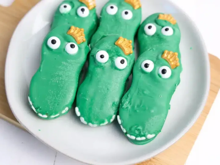Green chocolate-dipped Nutter Butter alligators with candy eyes and gold crowns on a white plate, fun safari animal party food ideas
