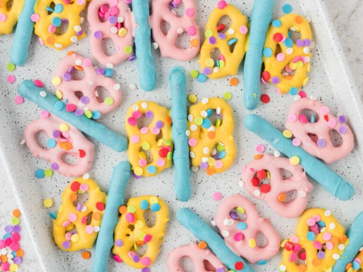 Colorful pink and yellow chocolate-dipped pretzels with blue pretzel sticks and rainbow confetti sprinkles on a white tray — a fun fairy party food idea.