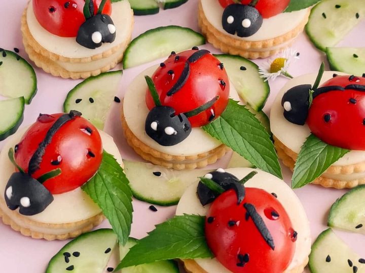 Caprese ladybug appetizers made with cherry tomatoes and black olives on crackers with cucumber and mint leaves — an adorable savory fairy party food idea.