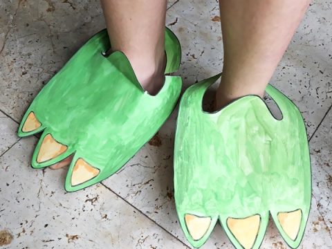 Child wearing large green painted cardboard dinosaur feet with yellow claws on a tiled floor