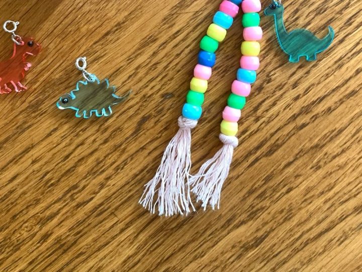 Colorful pony bead dinosaur keychain with a teal dino charm on a wooden surface beside a container of mixed beads