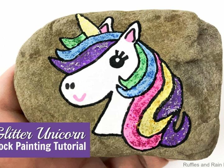 A hand holds a painted rock featuring a glitter rainbow unicorn and colorful mane as part of unicorn party crafts and activities