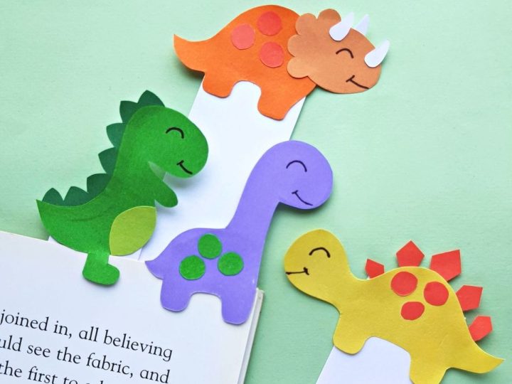 Four colorful paper dinosaur bookmarks in green, orange, purple, and yellow peeking out from an open book