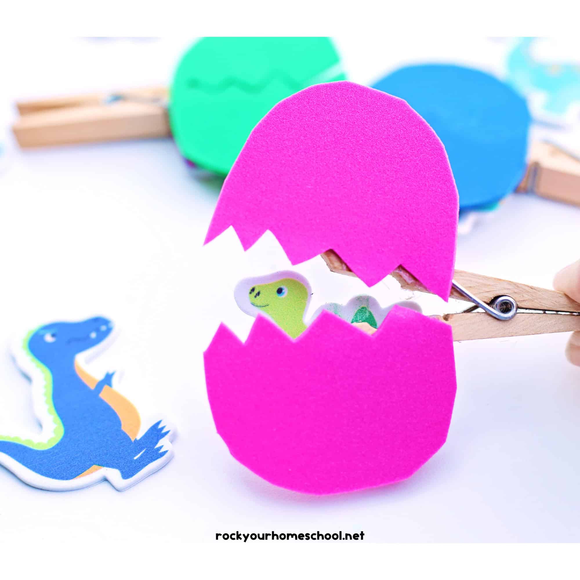 Bright pink foam hatching dinosaur egg craft opened with a clothespin revealing a baby dinosaur inside