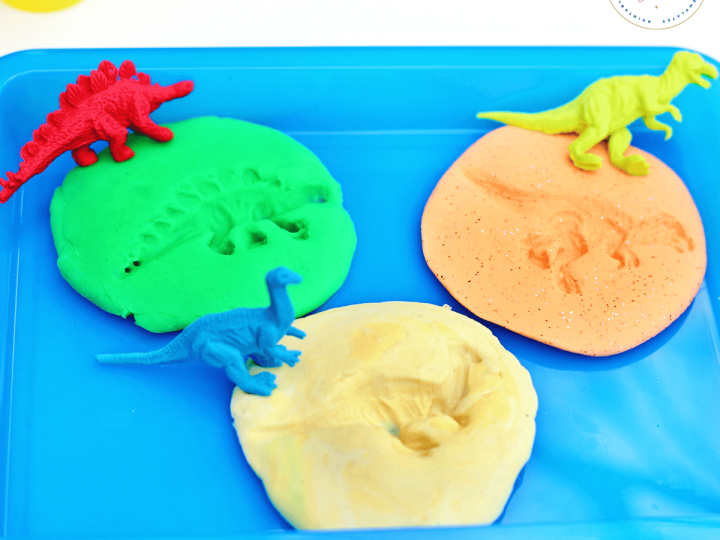 Green, orange, and yellow play dough rounds with dinosaur footprint impressions and small plastic dinosaur figurines on a blue tray