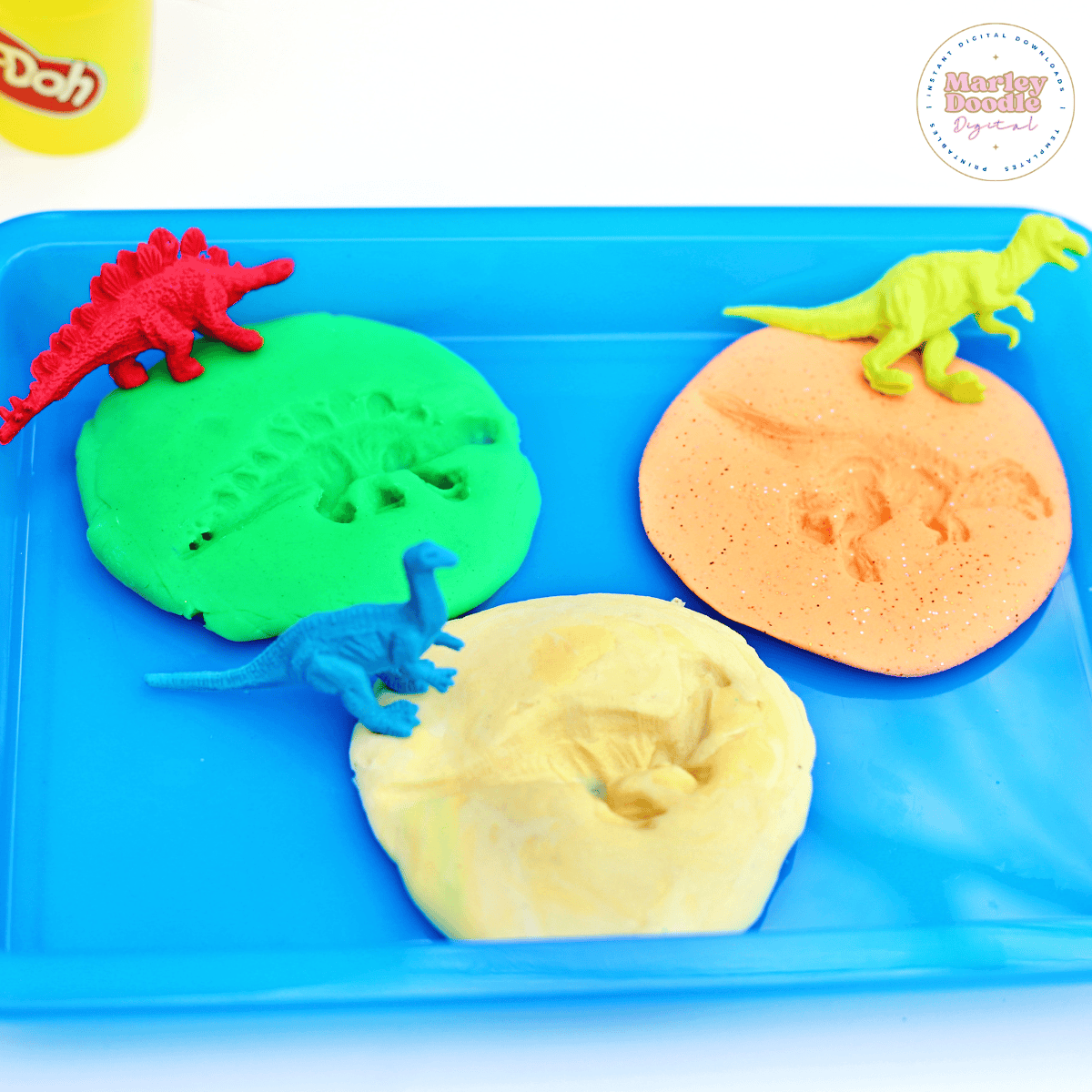 Green, orange, and yellow play dough rounds with dinosaur footprint impressions and small plastic dinosaur figurines on a blue tray