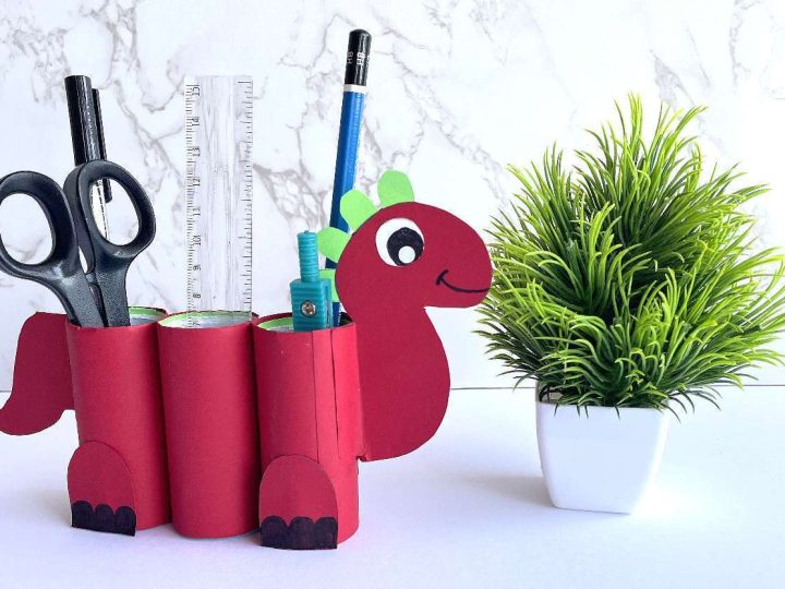 Red cardboard tube dinosaur pencil holder filled with scissors, ruler, and pencils on a white surface