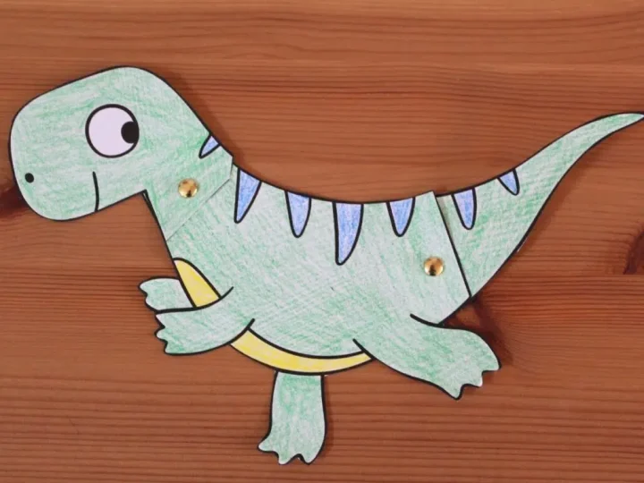 Green paper dinosaur craft with movable head and legs connected with brass fasteners on a wooden background
