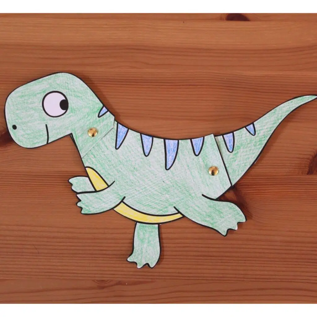 Green paper dinosaur craft with movable head and legs connected with brass fasteners on a wooden background