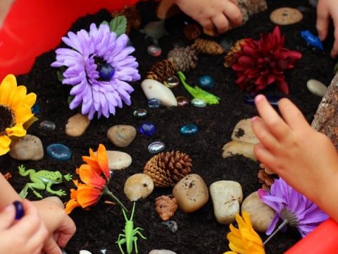 Children's hands exploring a fairy garden sensory bin filled with soil, flowers, rocks, and pine cones for fairy party crafts and activities.