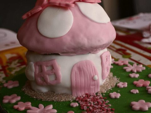 Pink fondant fairy toadstool birthday cake with a tiny door, flower sprinkles, and birthday candles — a magical fairy party food centerpiece for kids.