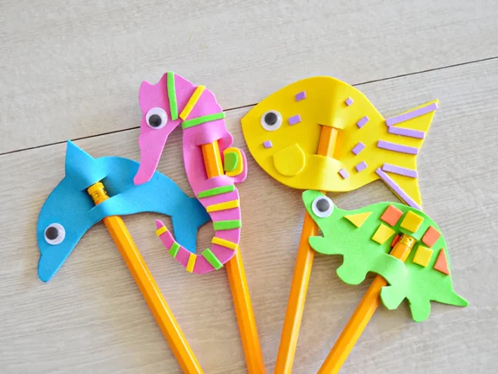 Four foam ocean animal pencil toppers shaped like a dolphin, seahorse, fish, and turtle on a wooden surface