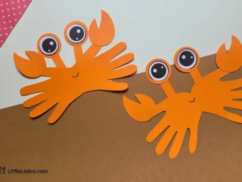 Two orange paper handprint crab crafts with googly eyes and paper claws on a two-toned beige background