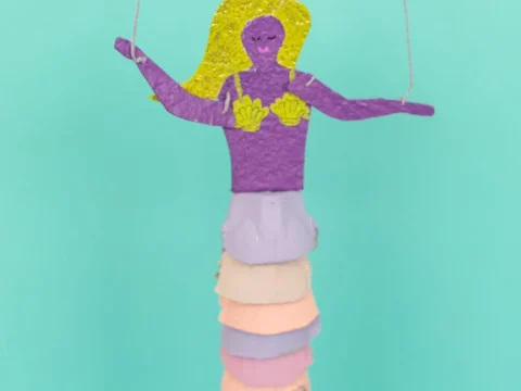 A handmade purple and yellow mermaid marionette toy with a pastel ombre tail hanging against a teal background