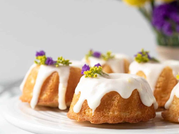 Mini glazed bundt cakes topped with drizzled white icing and tiny purple edible flowers on a white cake stand — an elegant fairy party food dessert idea.