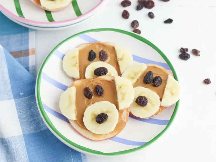 Apple slices topped with peanut butter, banana rounds, and raisin eyes arranged as monkey faces, a cute safari animal party food idea