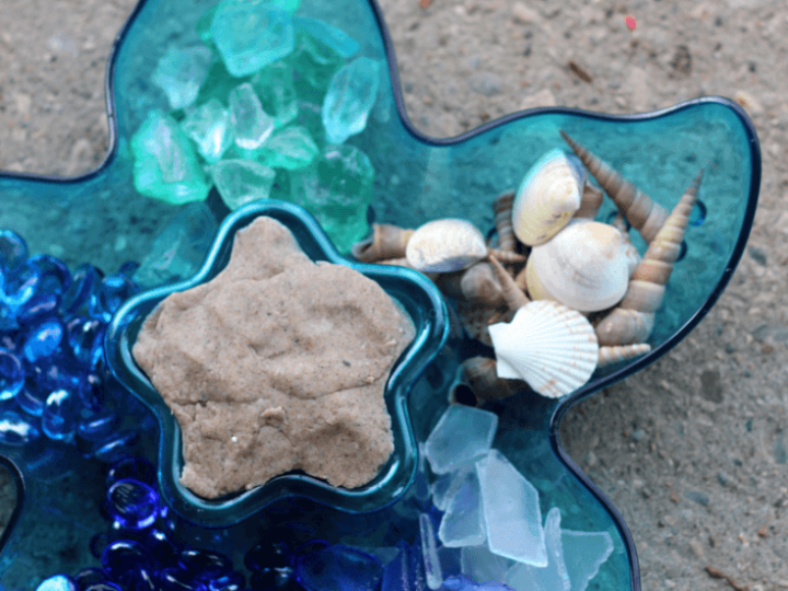 A blue starfish-shaped tray holding sandy play dough, seashells, sea glass, and blue gems for an ocean sensory activity