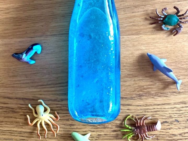 A blue sensory ocean bottle surrounded by small plastic ocean animal figurines on a wooden surface