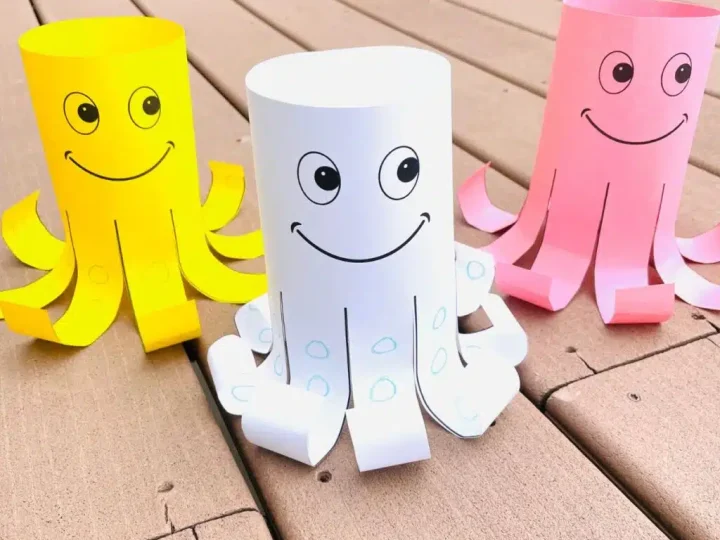 Three smiling paper tube octopus crafts in yellow, white, and pink with curled tentacles displayed on a wooden deck