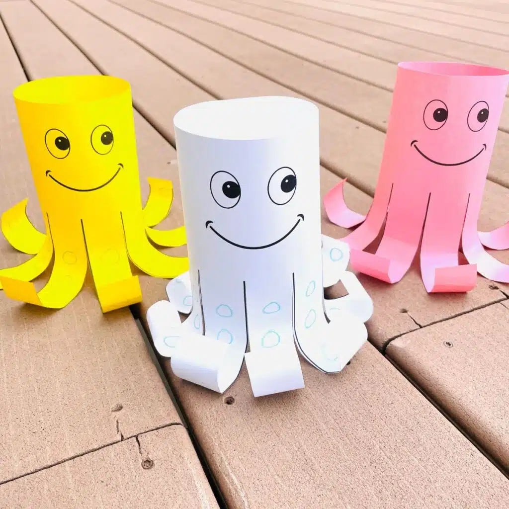 Three smiling paper tube octopus crafts in yellow, white, and pink with curled tentacles displayed on a wooden deck