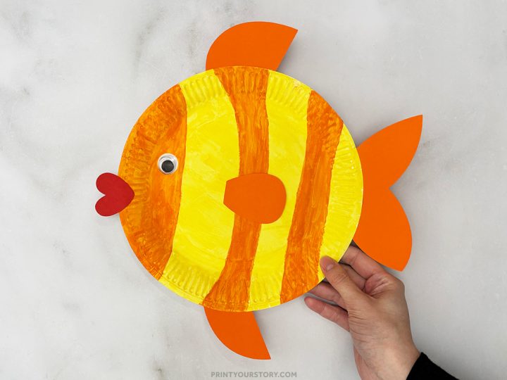 A hand holding a yellow and orange striped paper plate fish craft with a googly eye and red heart mouth on a marble surface