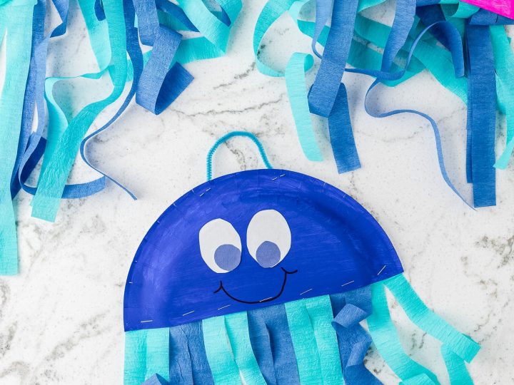 Three colorful paper plate jellyfish crafts in teal, pink, and blue with crepe paper tentacles on a marble surface