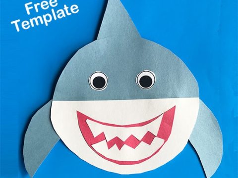 A smiling gray and white paper shark craft with googly eyes and pink teeth on a bright blue background