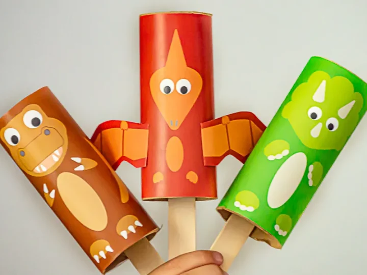 Three colorful paper tube dinosaur puppets in brown, red, and green held up by a child's hand
