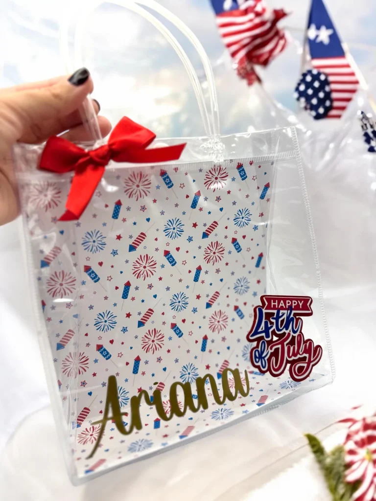 Clear plastic tote bag with fireworks and rocket print pattern, personalized gold name and Happy 4th of July sticker with red satin bow, festive 4th of July party favors.