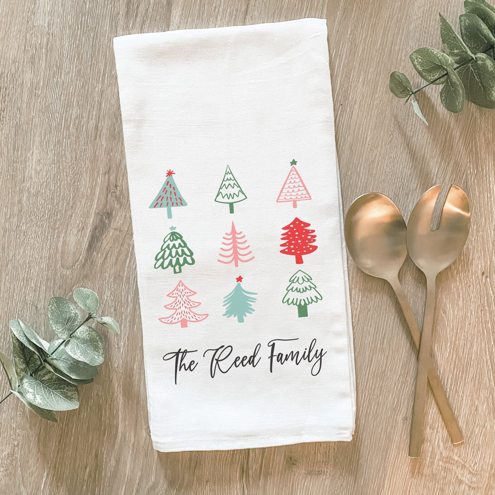 Personalized white tea towel with nine colorful Christmas tree designs and a custom family name โ a charming and practical Christmas party favor guests will love.