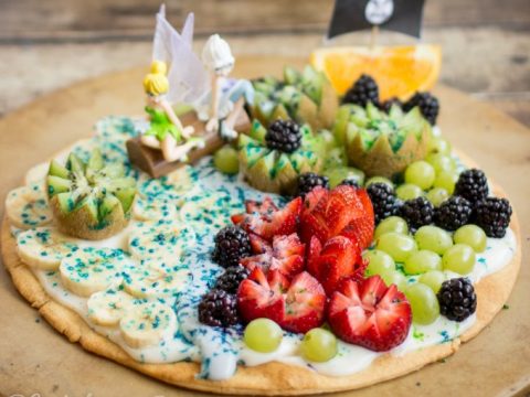 Pixie Hollow fruit pizza topped with strawberries, grapes, blackberries, kiwi, and banana with fairy figurines — a creative and healthy fairy party food centerpiece.