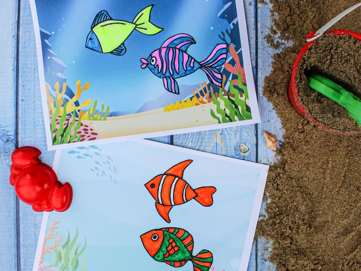 Two colorful printable underwater fish coloring pages displayed on a blue wooden surface beside a red sand bucket
