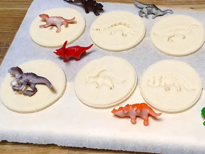 Six round salt dough discs with dinosaur fossil imprints displayed on parchment paper with small plastic dinosaur figurines