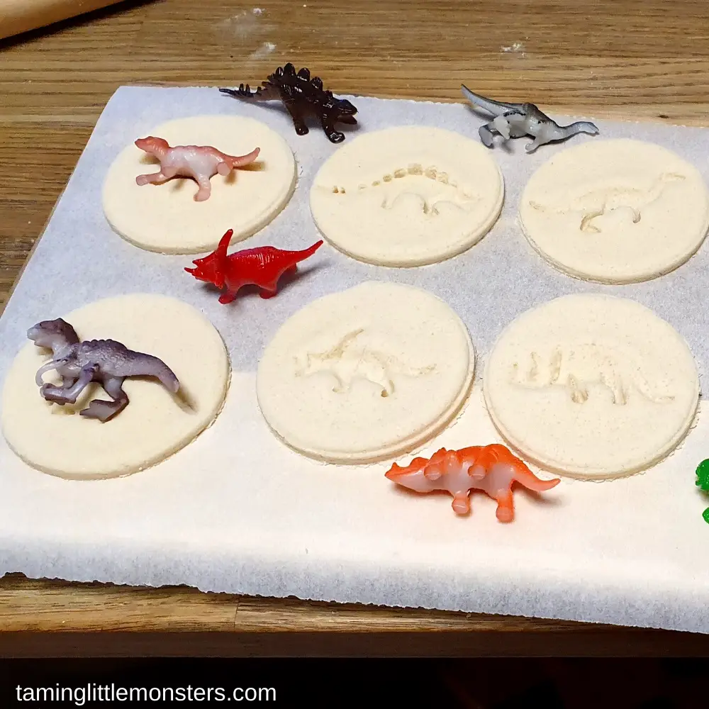 Six round salt dough discs with dinosaur fossil imprints displayed on parchment paper with small plastic dinosaur figurines