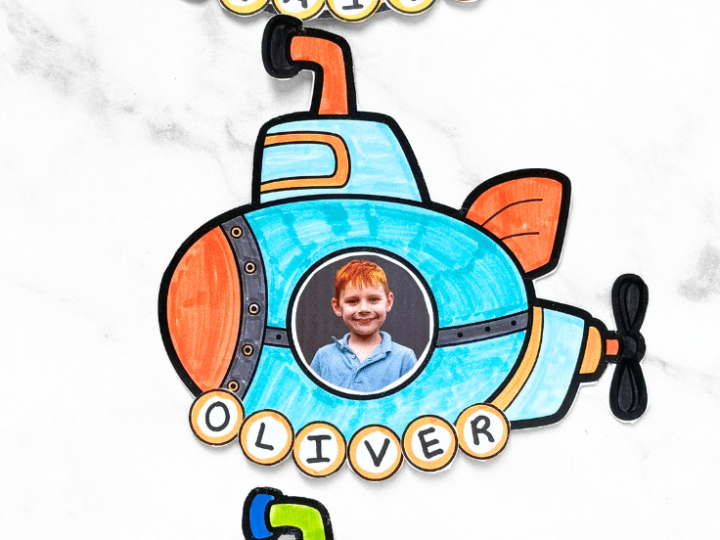 Three colorful printable submarine crafts with children's photos in the portholes displayed on a marble surface
