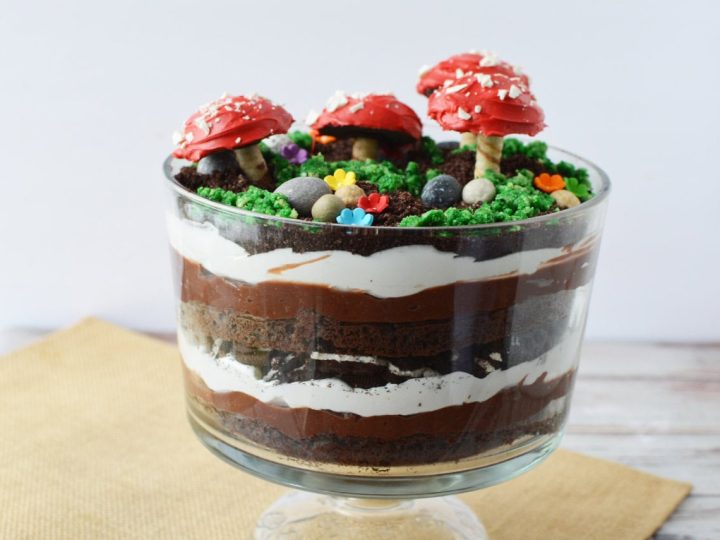 Layered chocolate trifle in a glass bowl topped with red toadstool decorations, green crumbs, and candy flowers — a showstopping fairy party food idea.
