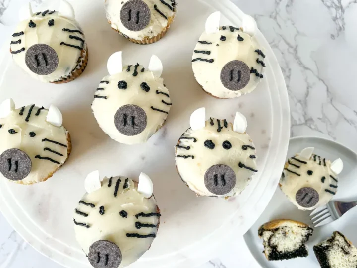White-frosted zebra face cupcakes with black stripe decorations and Oreo noses on a white cake stand, adorable safari animal party food ideas
