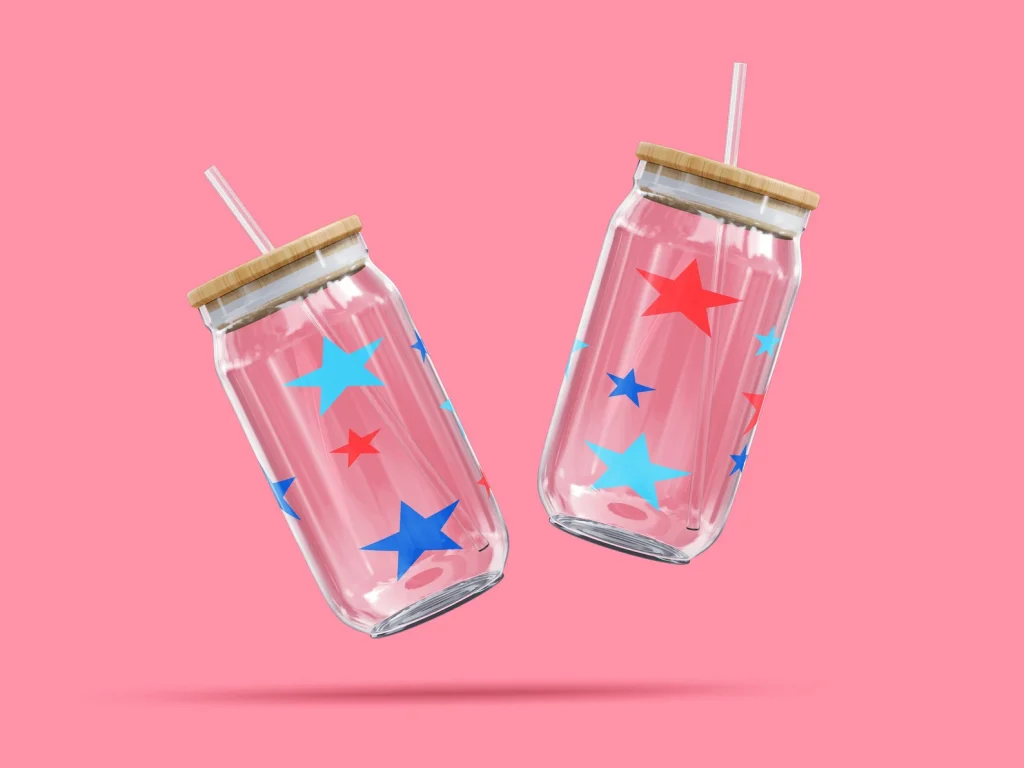 Two clear glass can-shaped tumblers with red and blue star designs and bamboo lids with straws on a pink background, trendy 4th of July party favors.