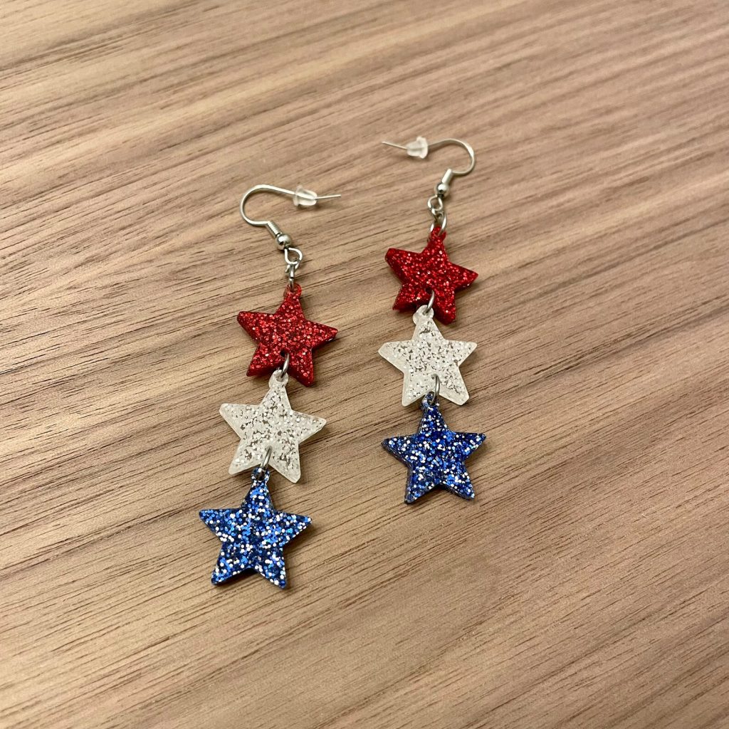 Two pairs of dangling triple glitter star earrings in red, white, and blue on a wooden surface, eye-catching 4th of July party favors.