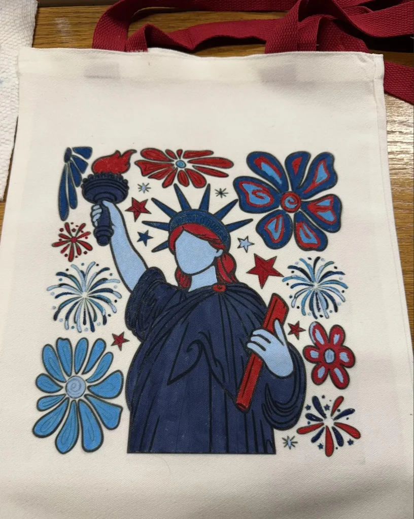 Hand-painted canvas tote bag featuring a colorful Statue of Liberty surrounded by red white and blue flowers and fireworks with red handles, artistic 4th of July party favors.