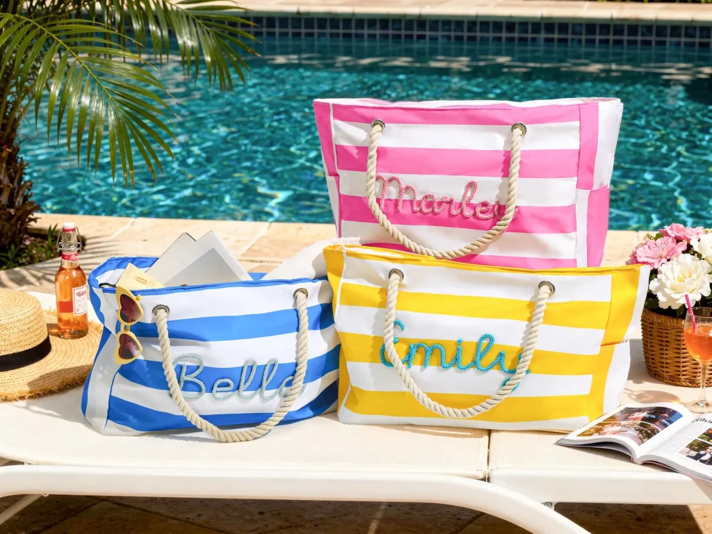 Three personalized striped beach tote bags in pink, blue, and yellow with rope handles beside a pool, charming pool party favors.