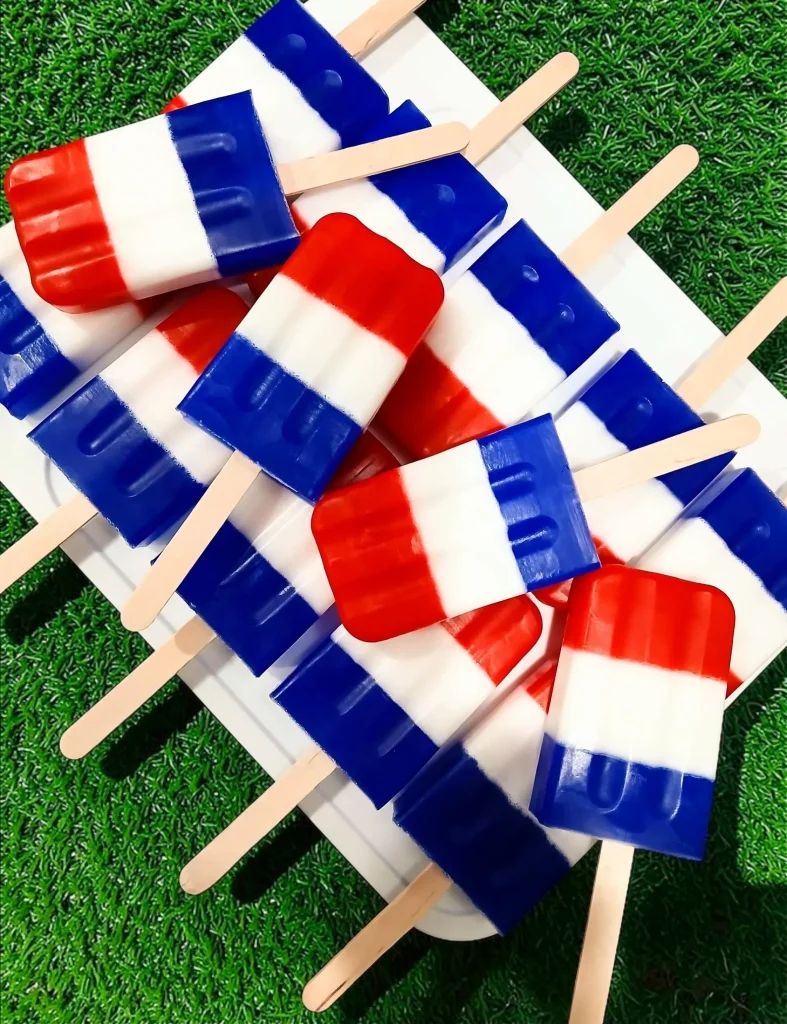Red white and blue striped popsicle-shaped soaps with wooden sticks piled on a white plate on green grass, fun and unique 4th of July party favors.