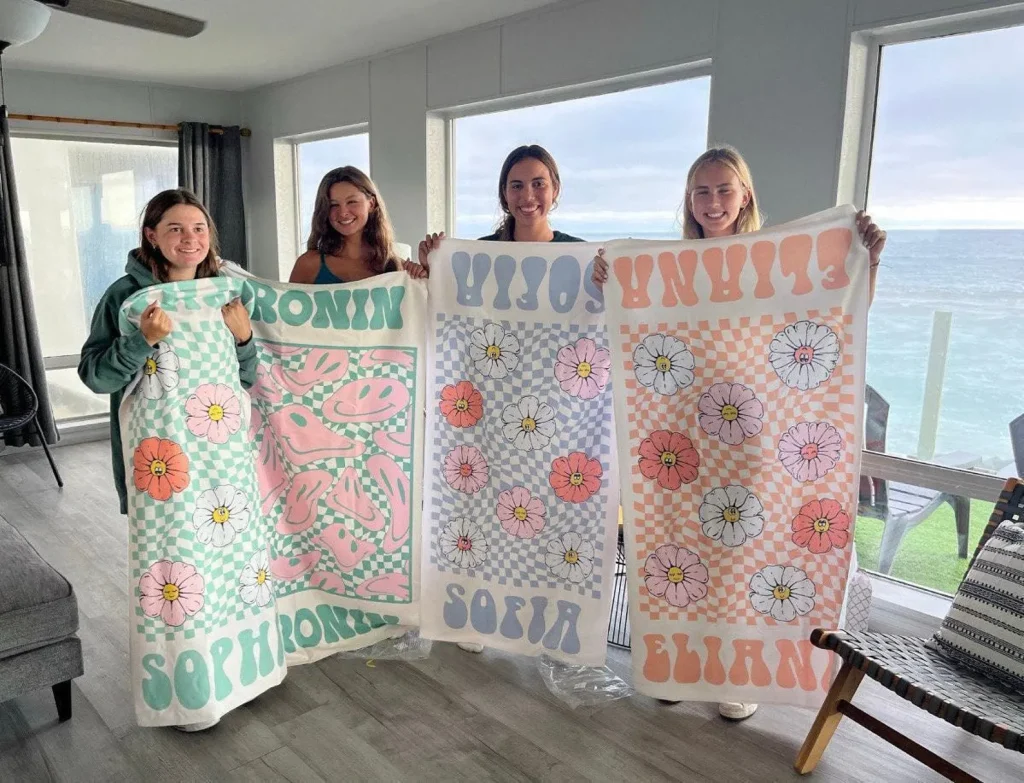 Four girls holding personalized retro checkerboard beach towels with daisy prints and custom names, perfect pool party favors for groups.