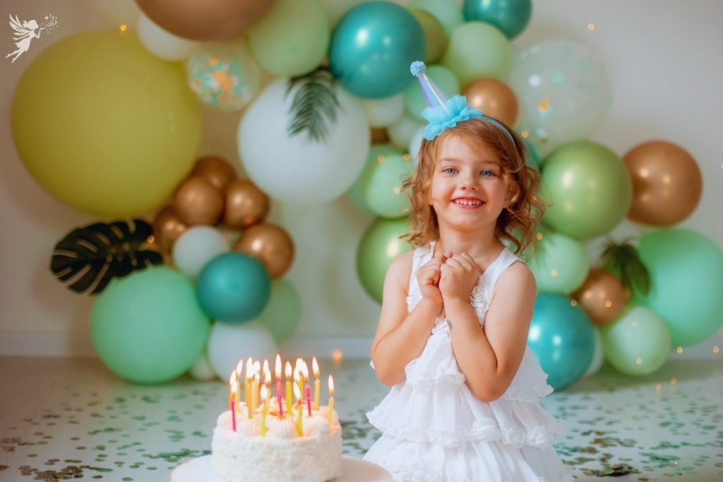little girl with a birthday cake full of candles smiling happily back drop green and gold balloons nature theme