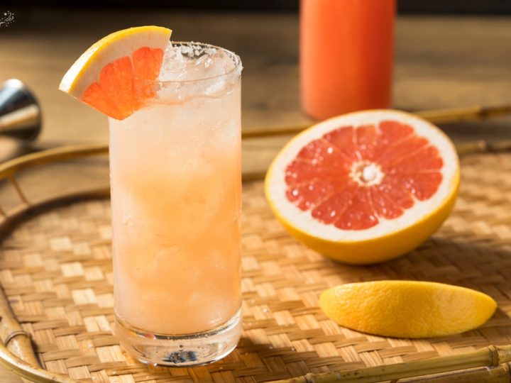 grapefruit paloma drink sitting on a ratten serving tray with a grapefruit slice and a grapefruit cut open